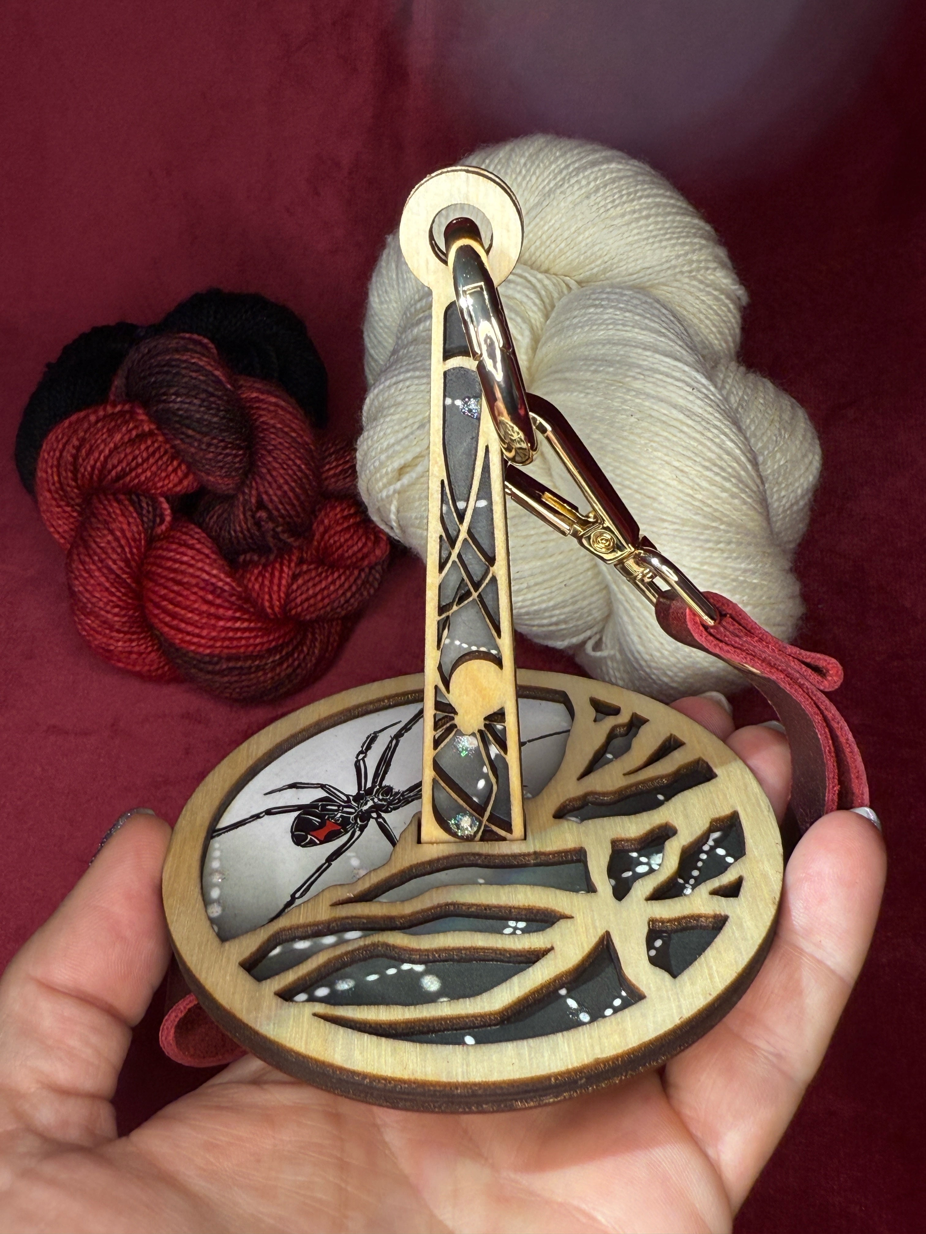 Decorative yarn holder with spider design held by a hand, against a red background with yarn.
