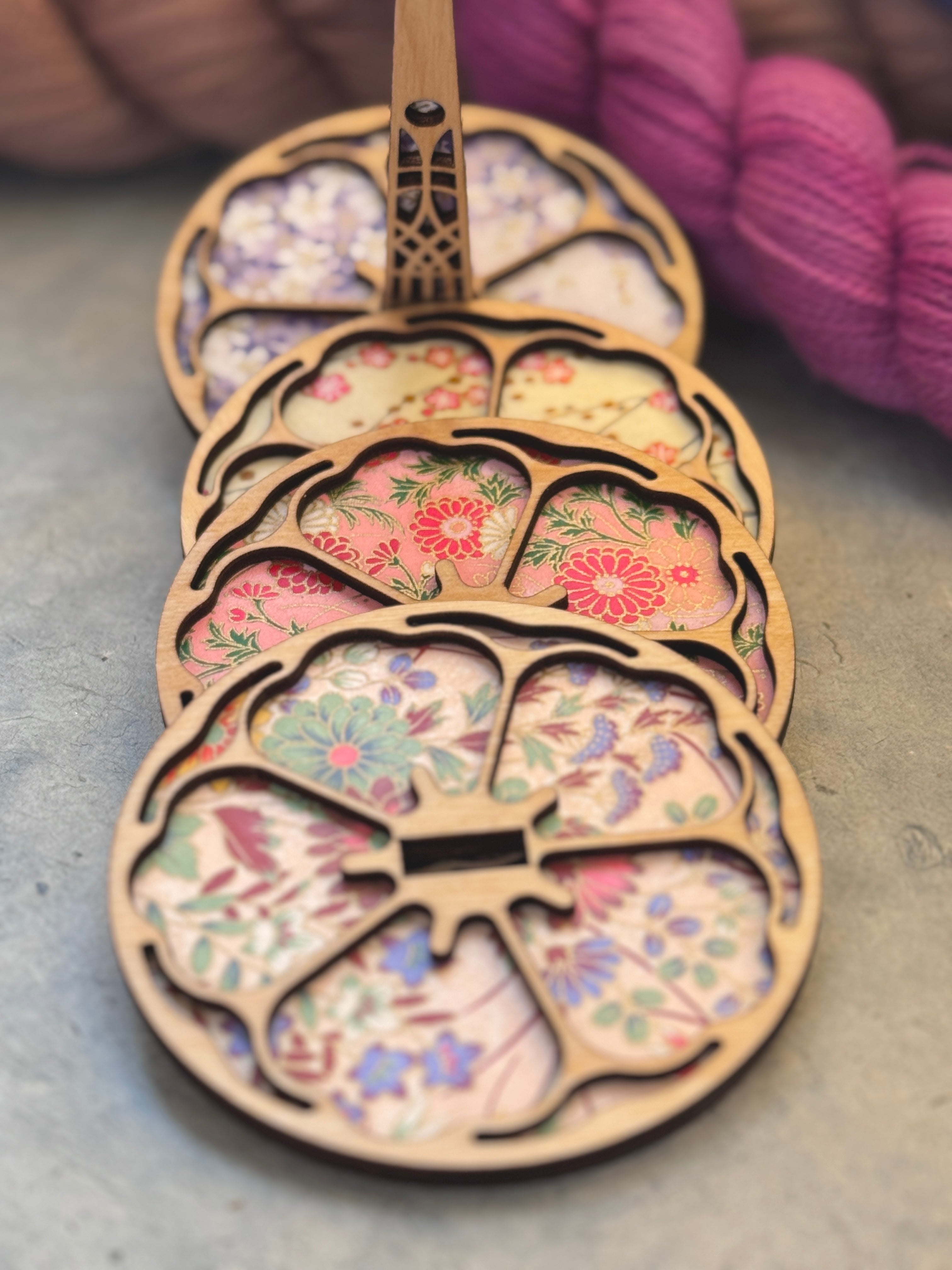 Yarn holder plates decorated with a laser cut-out of a flower in wood revealing different variations of decorative chiyogami paper designs.