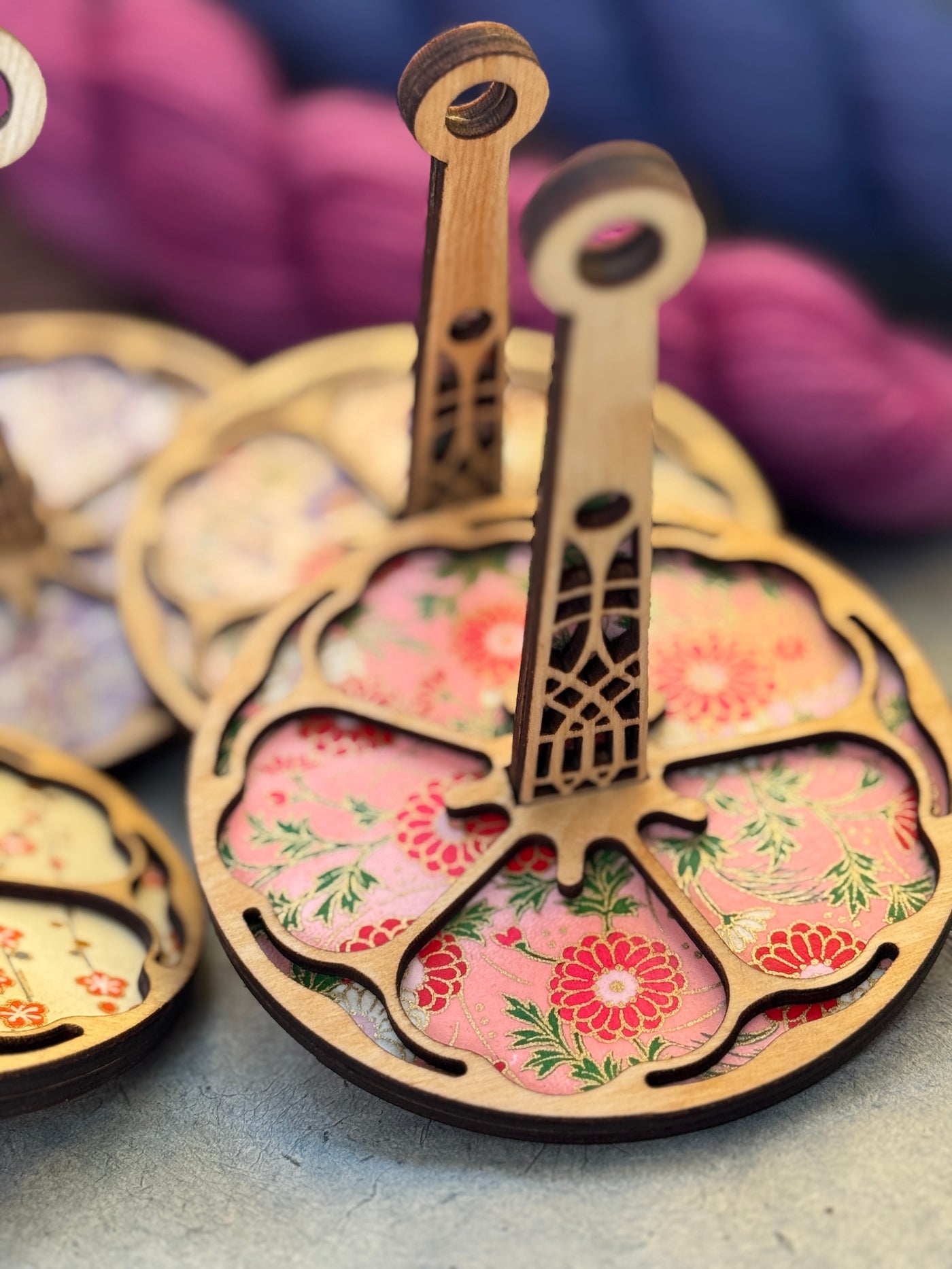 Wooden circular yarn holder with laser cut flower and a pink floral underlay on a textured gray surface.