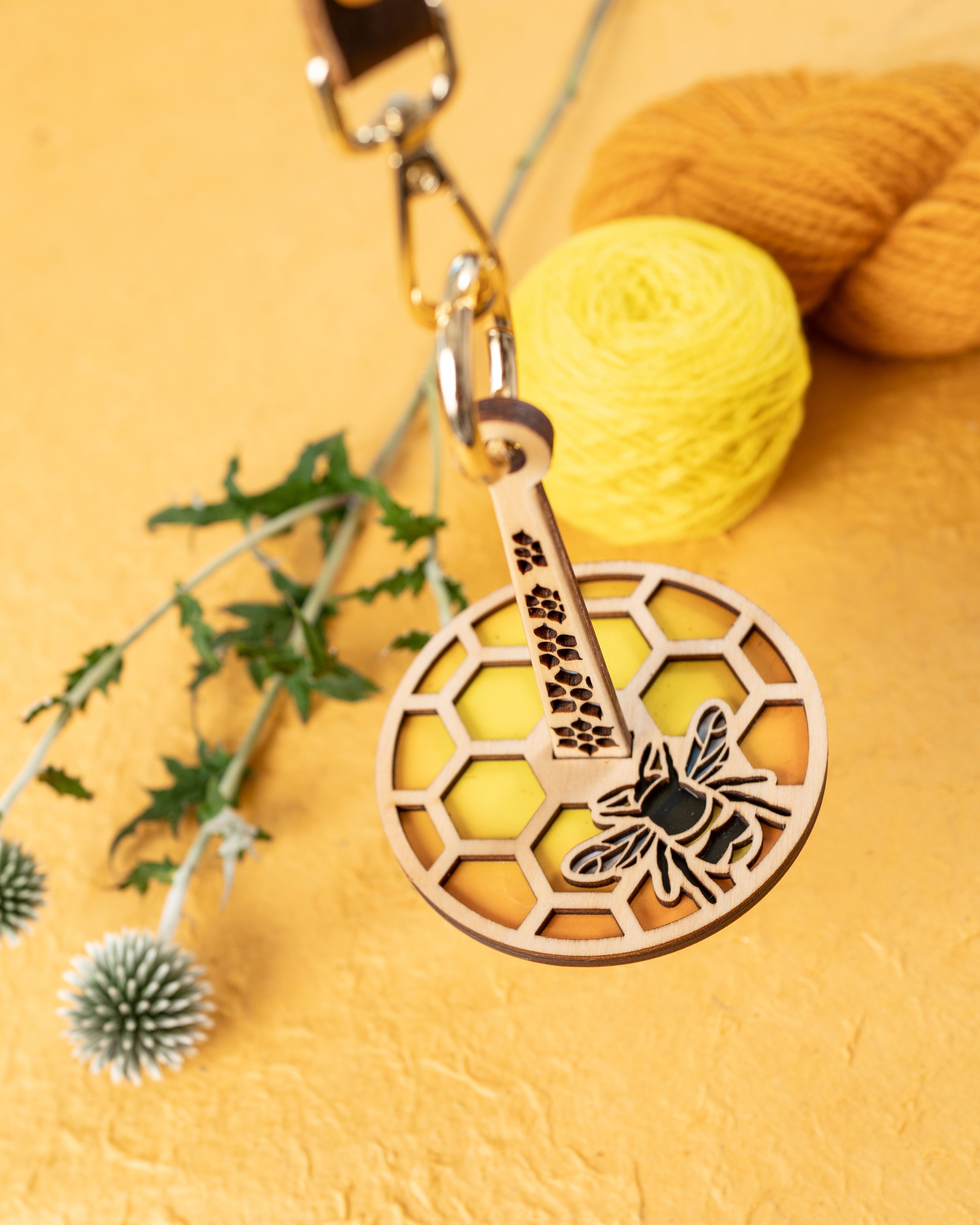A portable yarn holder with a honeycomb and bee design, attached to a leather strap, displayed alongside yarn balls and floral decor.