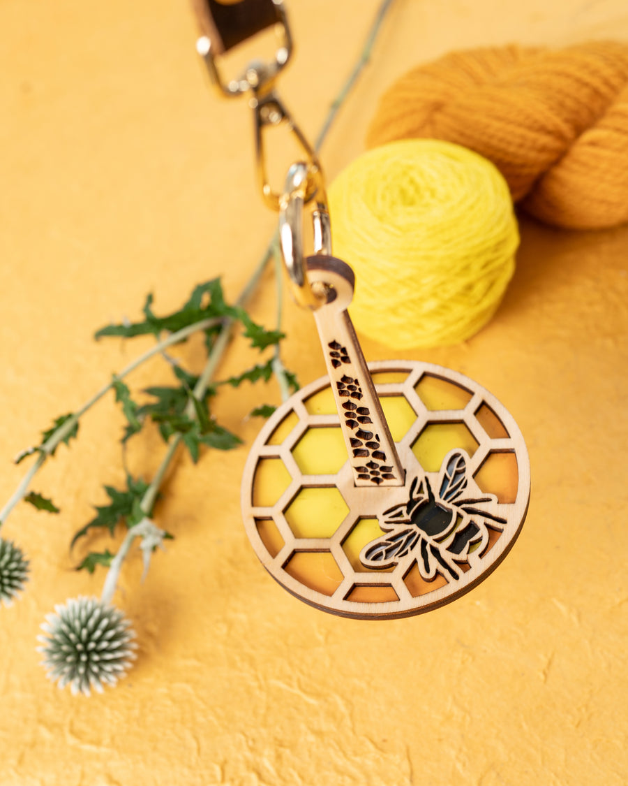 A portable yarn holder with a honeycomb and bee design, attached to a leather strap, displayed alongside yarn balls and floral decor.
