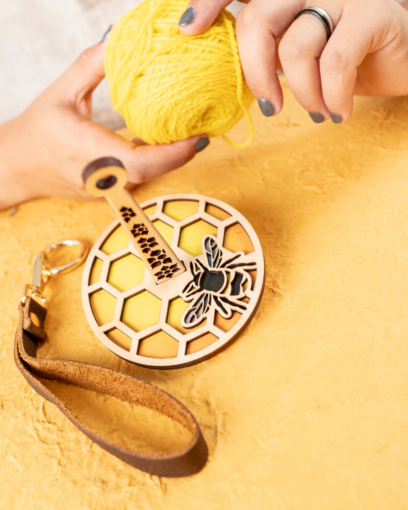 A wrist yarn holder sitting on a yellow surface. There is a honeybee design on the yarn holder. Behind the yarn holder there are hands holding a ball of yarn.