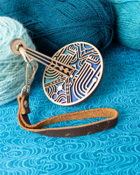 A decorative portable yarn holder with a blue art deco inspired design connected to a leather wrist strap to be used for knitting and crochet yarn management.