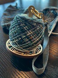 A wrist yarn holder sitting on a black wood table holding a ball of variegated blue and white yarn attached to a knitting project.