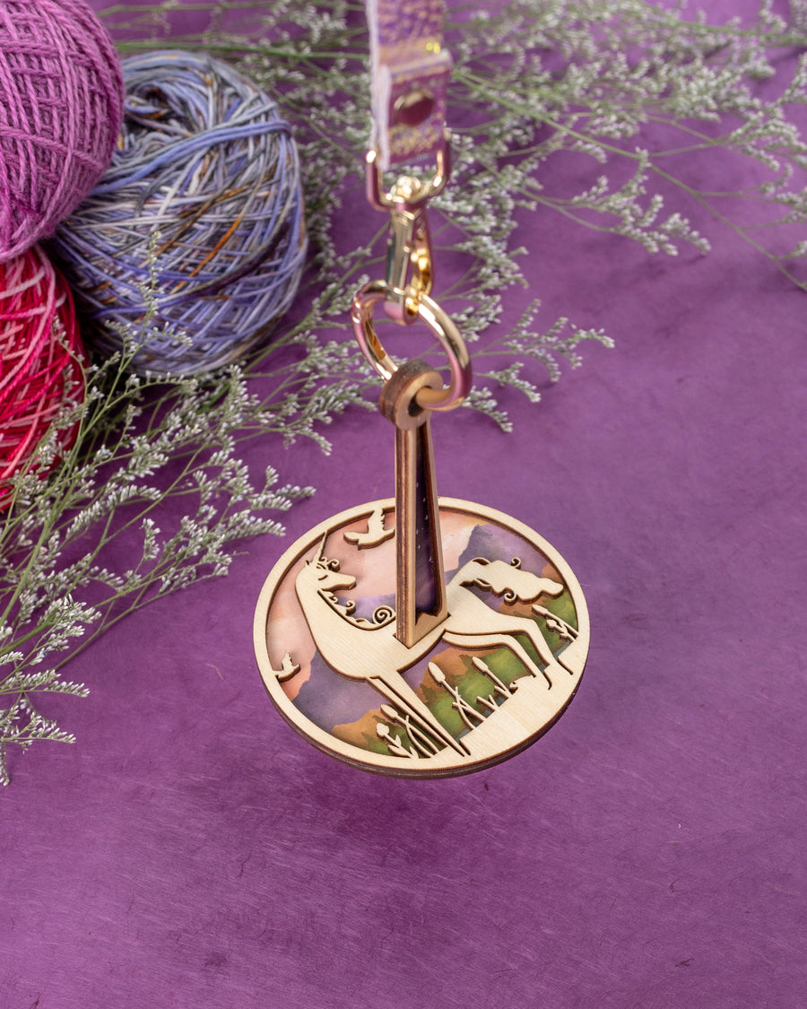 A wooden Mini Minder yarn holder with a unicorn pattern, attached to an iridescent leather strap, displayed on a purple background with yarn balls around it.