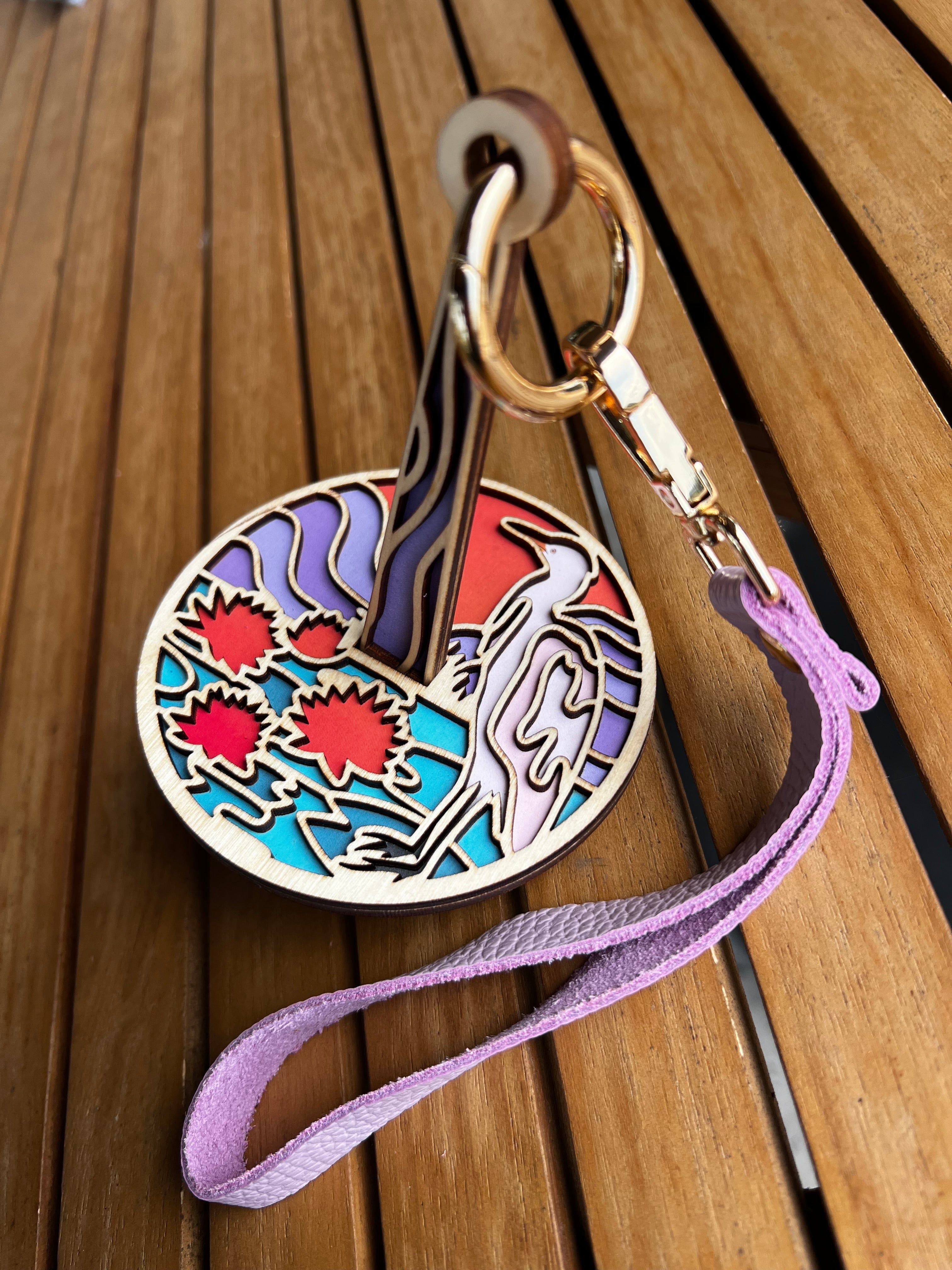 A laser cut wood yarn holder featuring a design of a crane like bird standing on a lily pad at sunset. The yarn ball holder is attached to a lavender leather strap and is displayed on a slatted wood surface.