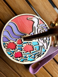 A closeup up the plate of a circular yarn holder. A laser cut wood design displays a crane like bird standing on a lily pad at sunset surrounded by purple mountains and crimson flowers. 
