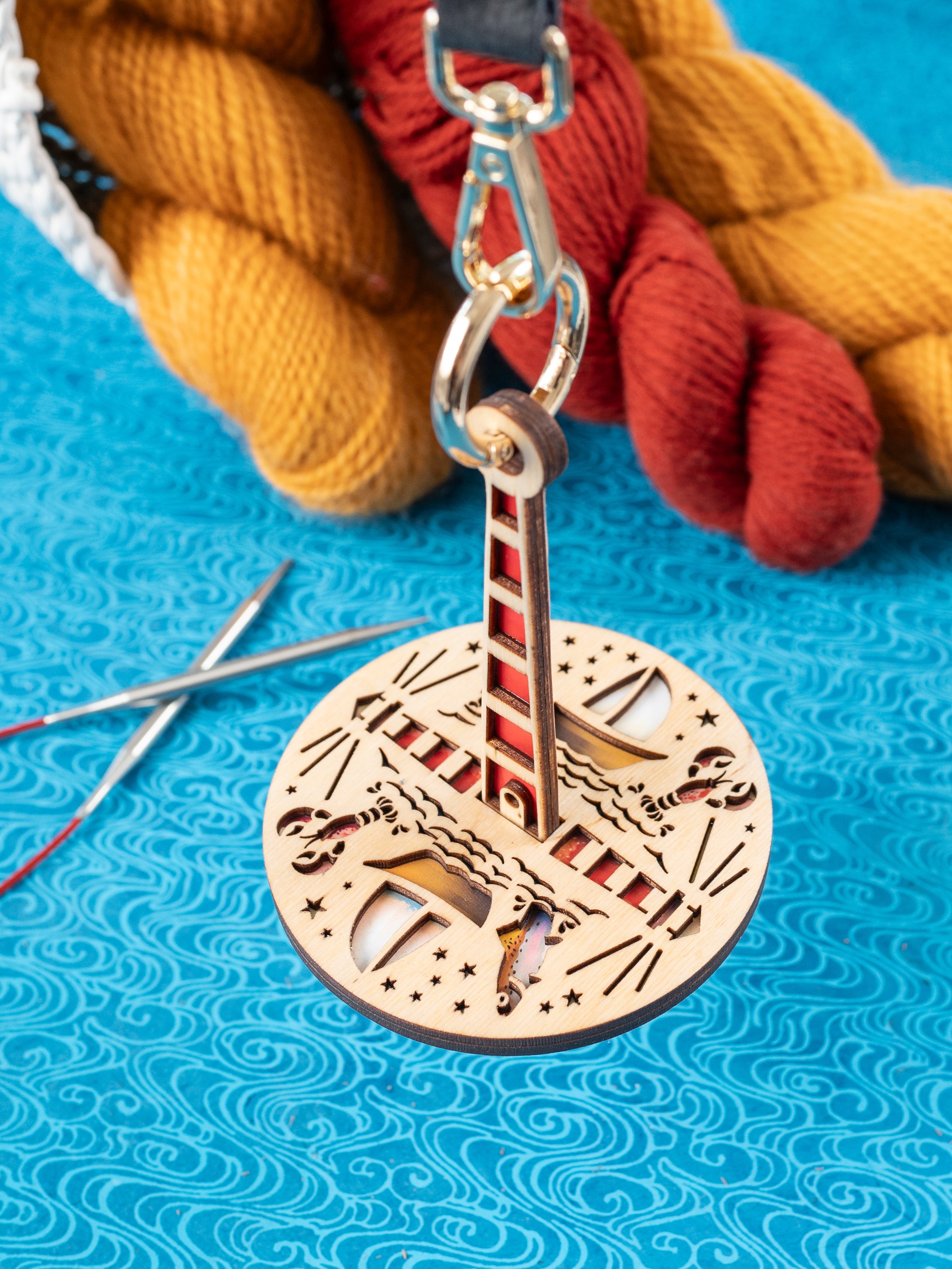 A wooden yarn minder featuring ocean-themed graphics, dangling from a leather strap, on a blue textured surface with yarn in the background.