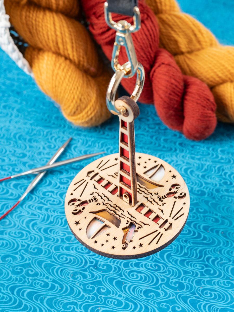 A wooden yarn minder featuring ocean-themed graphics, dangling from a leather strap, on a blue textured surface with yarn in the background.
