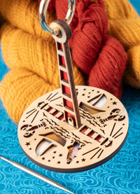 A wooden yarn ball holder featuring ocean-themed graphics, dangling metal ring, on a blue textured surface with yarn in the background.