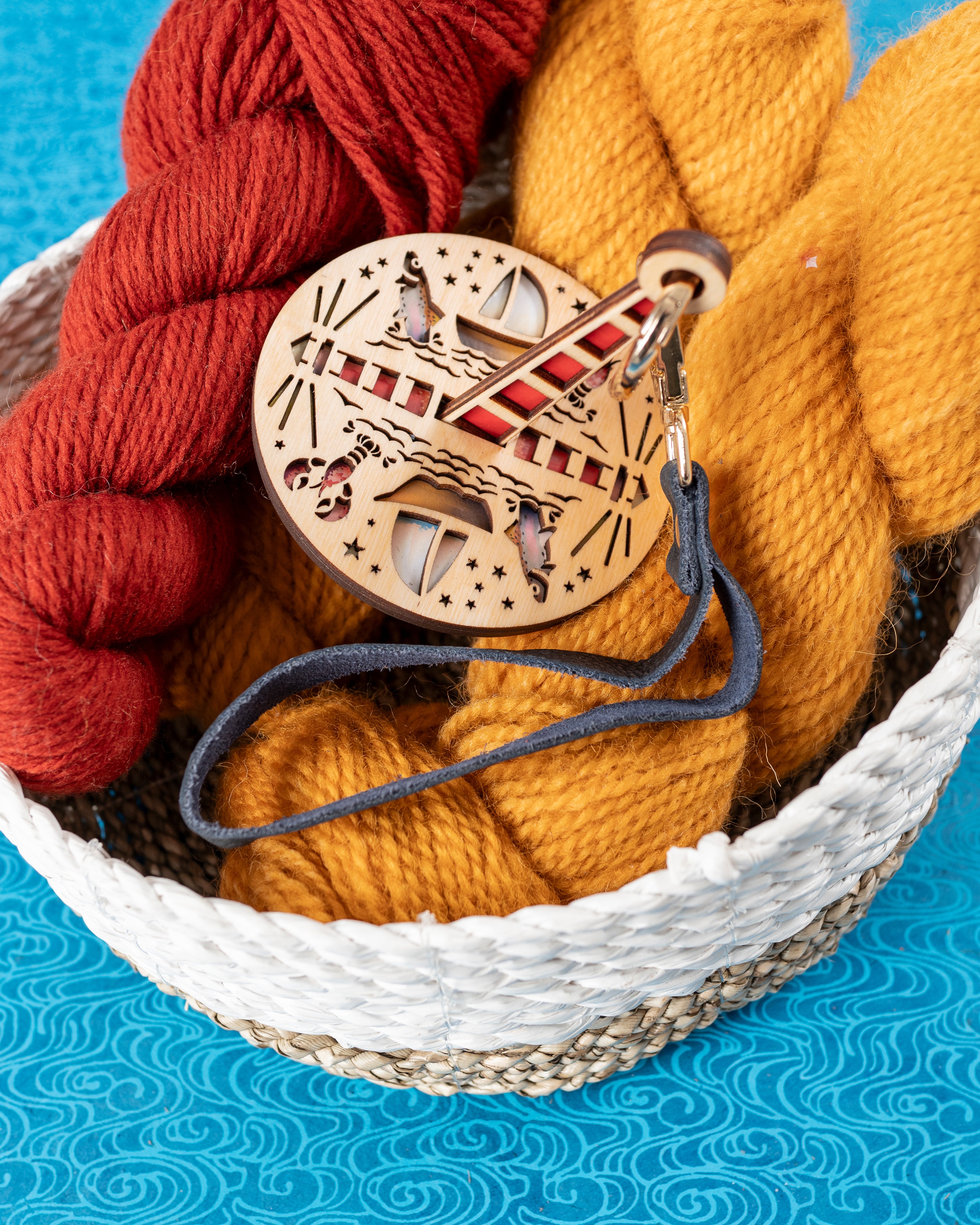 A wooden yarn holder featuring ocean-themed graphics, with an attached dark blue leather strap, sitting on top of a basket full of yarn.