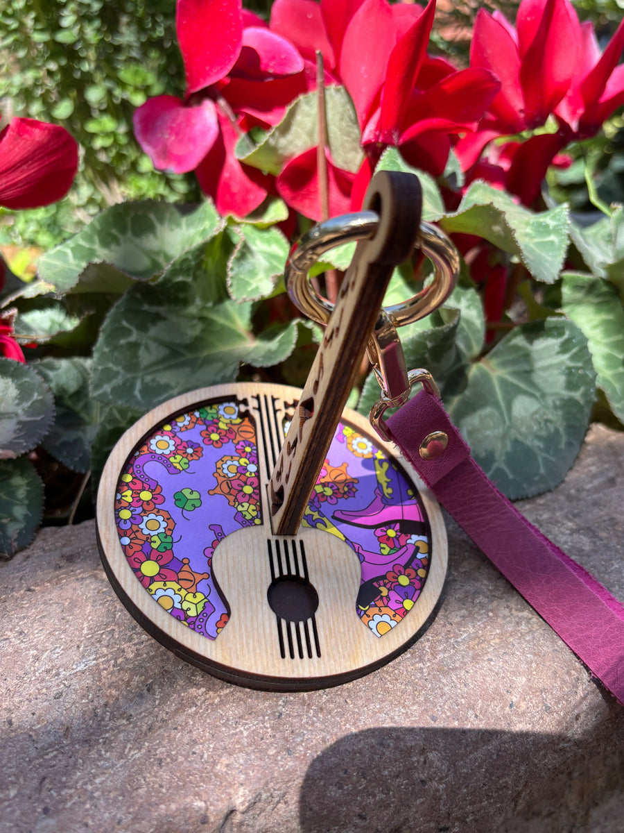 A portable yarn holder with a wrist strap, decorated with a colorful floral design and a guitar motif, placed on a rock beside red flowers.