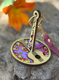 A colorful floral Mini Minder yarn holder with a guitar design and wrist strap, resting on a rock beside vibrant orange flower.