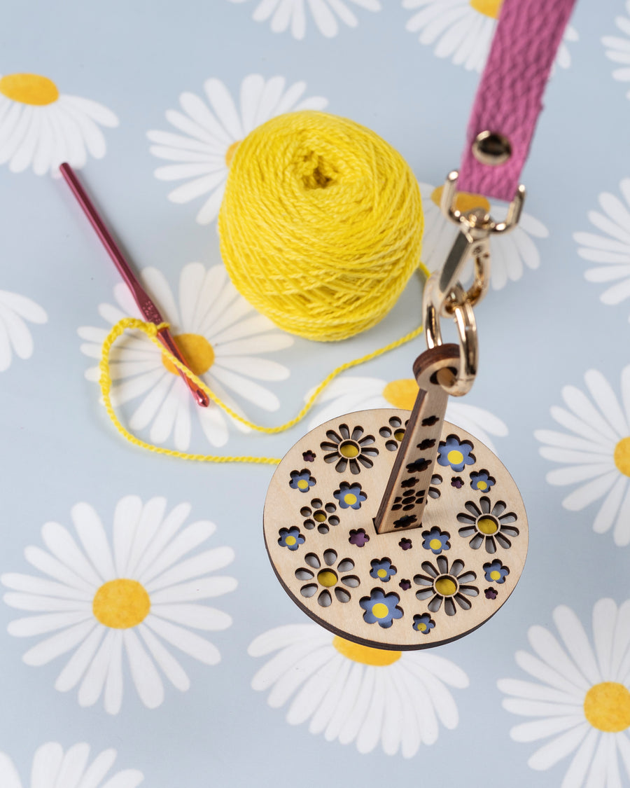 A portable yarn holder with a floral pattern, resembling a wrist accessory, accompanied by a ball of yellow yarn and a crochet hook, displayed on a fabric with daisy prints.