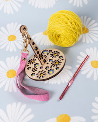 A portable yarn minder with a floral pattern, with an attached pink leather strap, accompanied by a ball of yellow yarn and a crochet hook, displayed on a fabric with daisy prints.