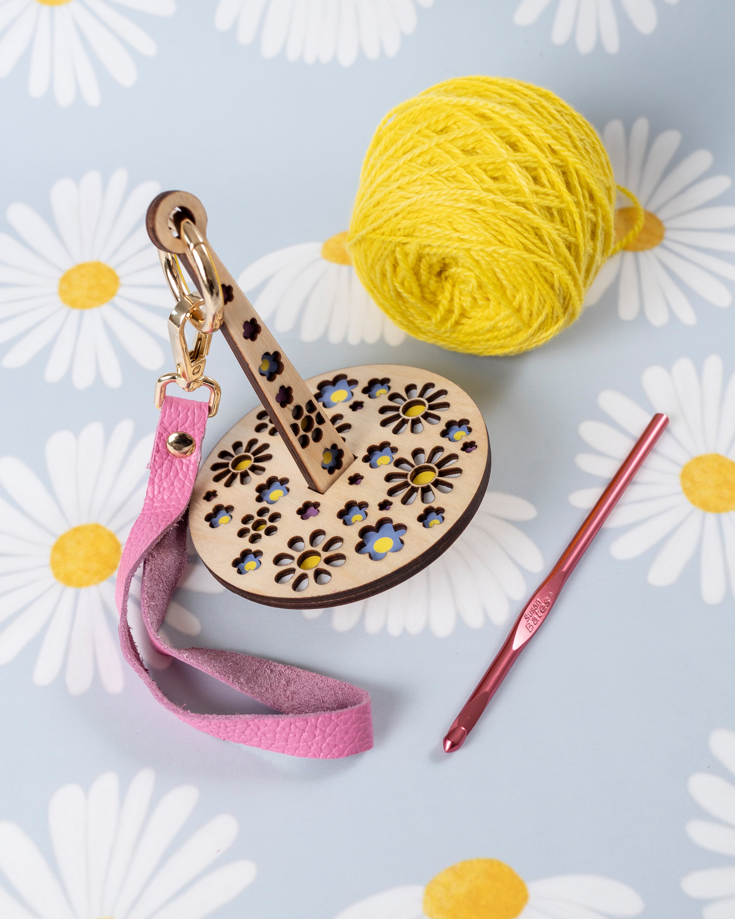 A portable yarn minder with a floral pattern, with an attached pink leather strap, accompanied by a ball of yellow yarn and a crochet hook, displayed on a fabric with daisy prints.