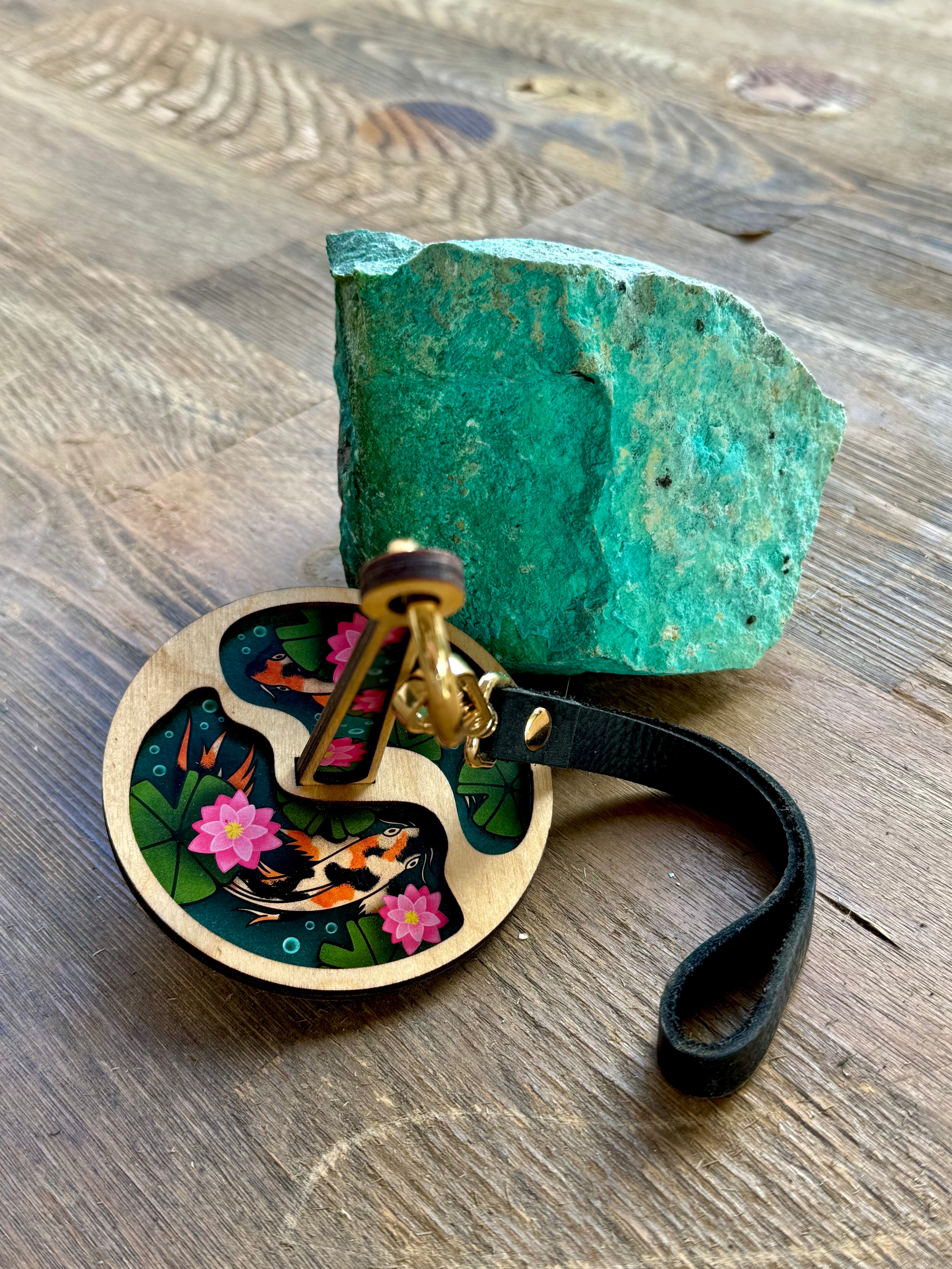 A portable yarn minder decorated with two koi fish swimming among green lily pads and pink lotuses, displayed beside a turquoise rock on a wooden surface.