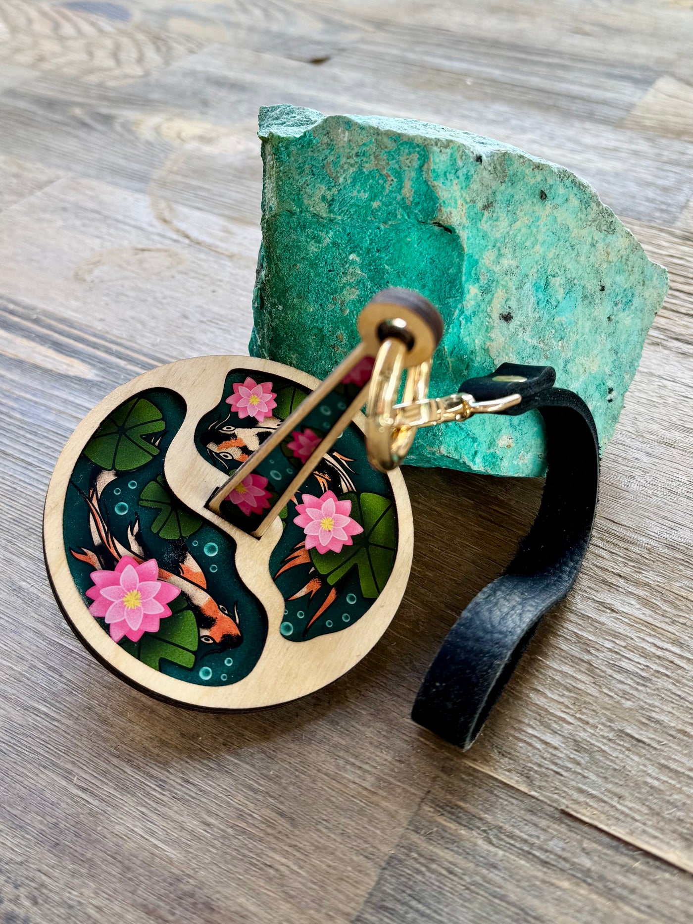 A wrist yarn holder featuring a design of two koi fish swimming in a pond with green lily pads and pink lotuses, next to a turquoise colored rock and sitting on a wooden surface.