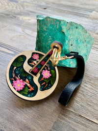 A wrist yarn holder featuring a design of two koi fish swimming in a pond with green lily pads and pink lotuses, next to a turquoise colored rock and sitting on a wooden surface.