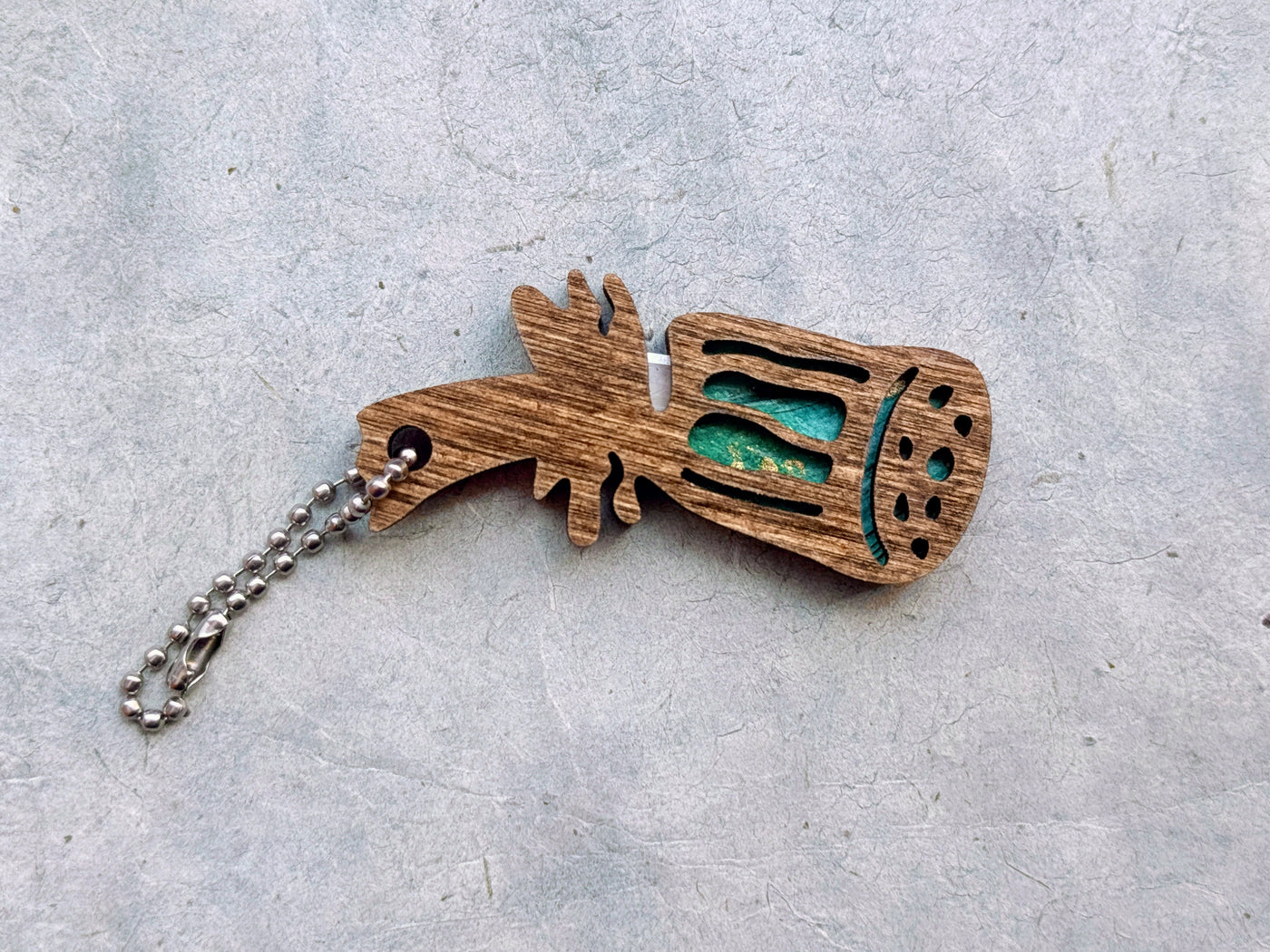 A lotus-head-shaped yarn cutter made from dark-stained wood and turquoise paper, with a ball chain attached, sitting on a textured light gray background.