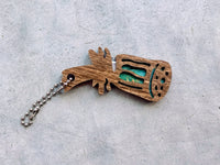 A lotus-head-shaped yarn cutter made from dark-stained wood and turquoise paper, with a ball chain attached, sitting on a textured light gray background.
