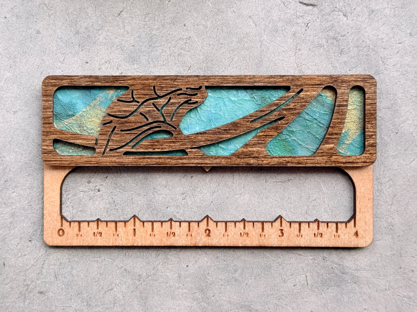 A gauge swatch made from dark-stained wood and turquoise paper, with a laser-cut flower design. There are numbers etched on the bottom indicating inch measurements. It's sitting on a textured light gray background.