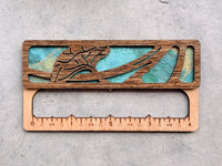 A gauge swatch made from dark-stained wood and turquoise paper, with a laser-cut flower design. There are numbers etched on the bottom indicating inch measurements. It's sitting on a textured light gray background.
