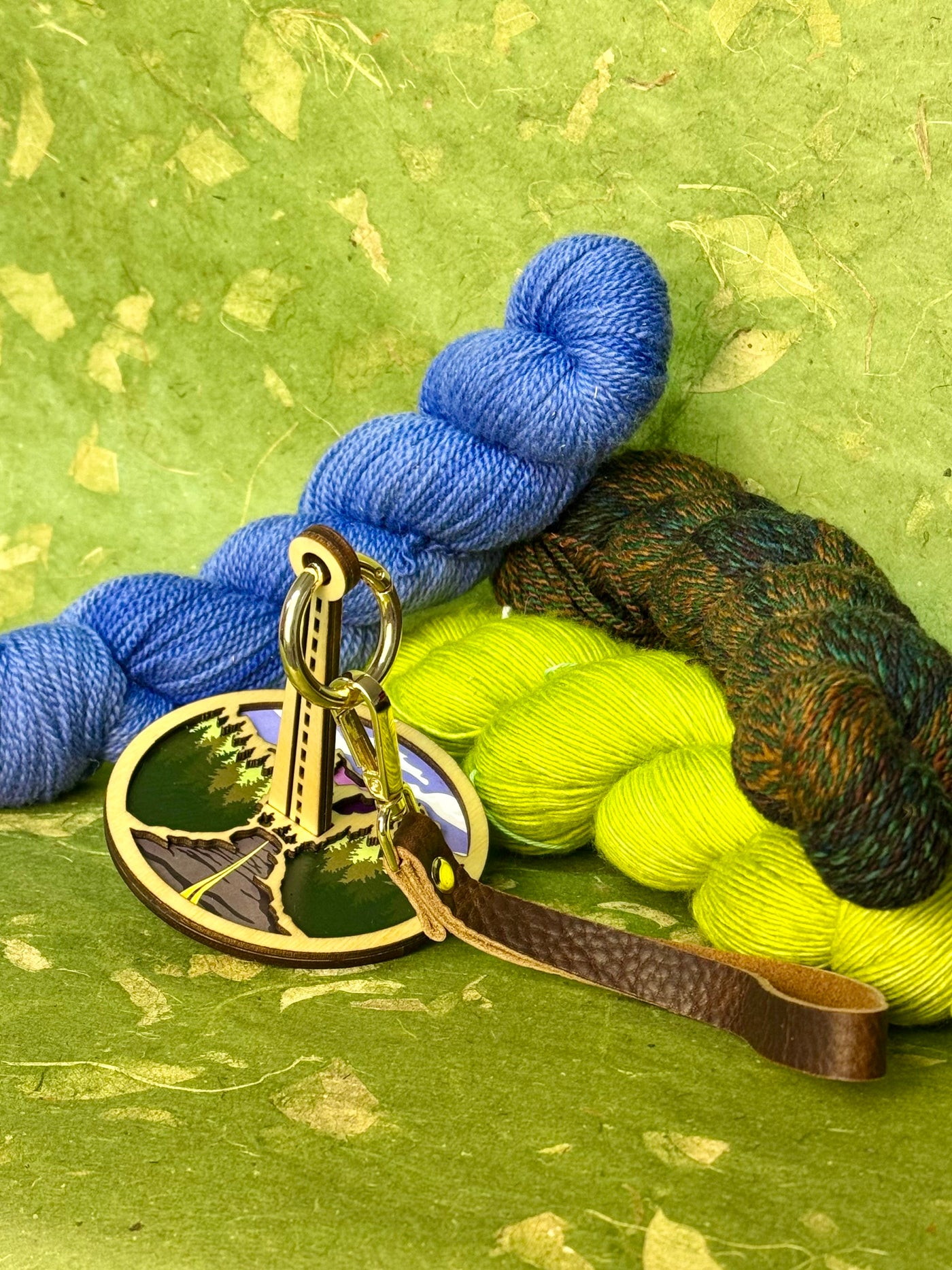Mountain‑themed portable yarn holder with a brown leather strap, photographed on a green background and framed by skeins of yarn.