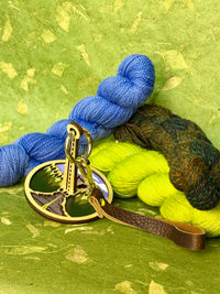Mountain‑themed portable yarn holder with a brown leather strap, photographed on a green background and framed by skeins of yarn.