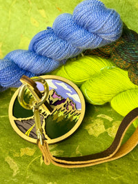 Close up of a travel‑friendly yarn holder showcasing a mountain cutout design and brown leather strap, set on a green surface with yarn skeins.