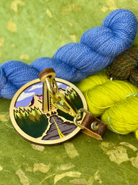 Portable yarn holder featuring a laser cut mountain scene. It has a brow leather strap attach to is and is sitting on a green background surrounded by yarn.